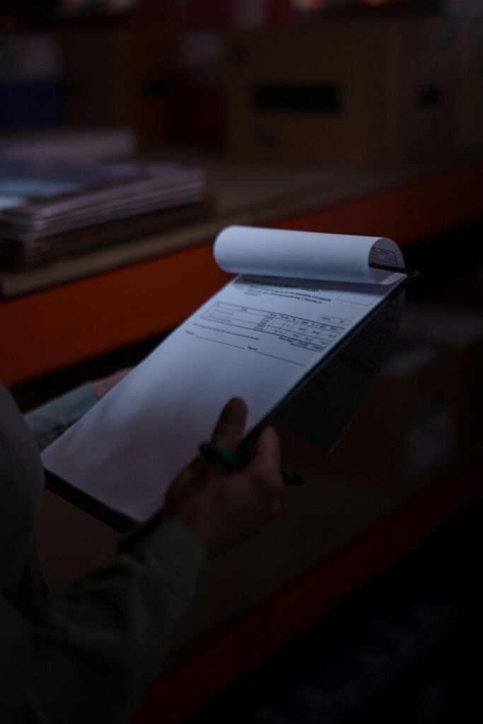 pexels-photo-7018654-7018654 Silhouette of a person holding a clipboard with documents in a dimly lit warehouse.