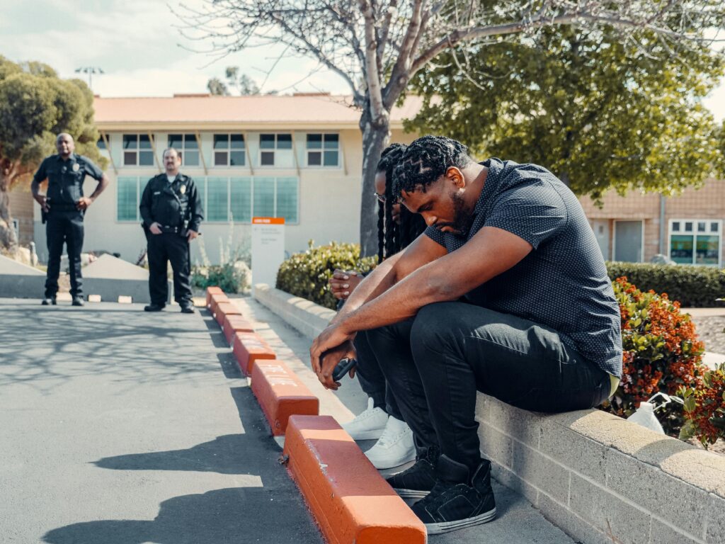 pexels-photo-7714865-7714865 Police officers observing two adults sitting on a curb, possibly detained, in a sunny outdoor setting.